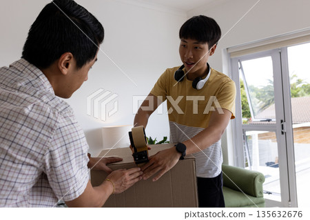 Father and son sealing cardboard box with tape dispenser at home, smiling together 135632676