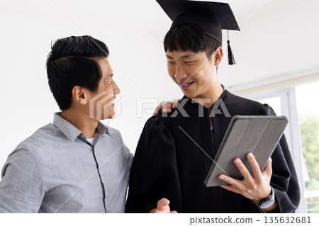 Proud father congratulating son in graduation gown holding tablet at home Proud father congratulating son in graduation gown holding tablet at home 135632681
