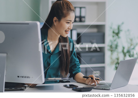 Determined asian businesswoman works late in a modern office, confidently analyzing data on her laptop, embodying success and professionalism with a smile Determined asian businesswoman works late in a modern office, confidently analyzing data on her laptop, embodying success and professionalism with a smile 135632856