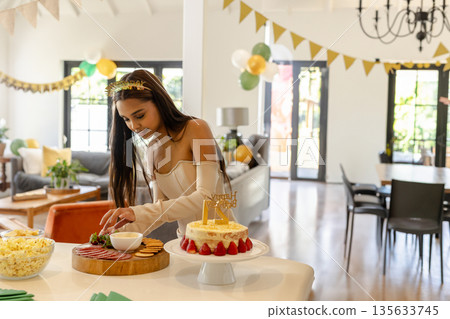 South Asian woman arranging wooden platter beside 21 cake under chandelier with balloons at home 135633745