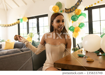 Woman wearing dress sitting at wooden table in living room with Happy Birthday banner and balloons Woman wearing dress sitting at wooden table in living room with Happy Birthday banner and balloons 135633749