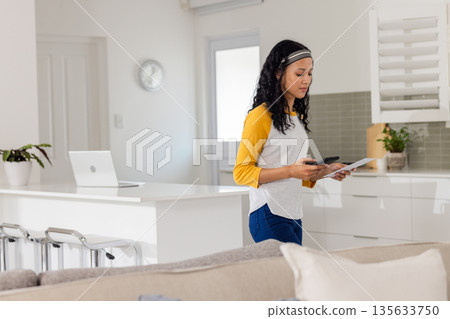 Teenage girl standing at kitchen island holding smartphone and paper with closed laptop, copy space Teenage girl standing at kitchen island holding smartphone and paper with closed laptop, copy space 135633750