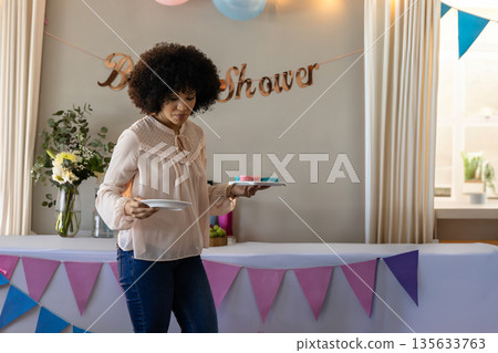 African American woman holding plates and cookies at home baby shower table, copy space 135633763