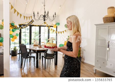 Female in early twenties holding birthday cake with gold 21 topper in dining room, copy space Female in early twenties holding birthday cake with gold 21 topper in dining room, copy space 135633765