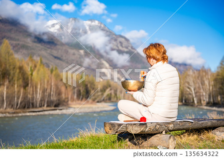 Woman playing a Tibetan singing bowl by a mountain river in Altai. Concept of sound healing, spiritual alignment, and deep connection with natural elements 135634322