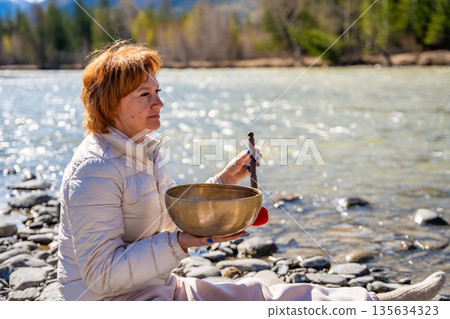 Woman performing a sound ritual with a Tibetan singing bowl by the Altai river. Concept of feminine energy, sacred vibration, and shamanic connection to the Earth Woman performing a sound ritual with a Tibetan singing bowl by the Altai river. Concept of feminine energy, sacred vibration, and shamanic connection to the Earth 135634323