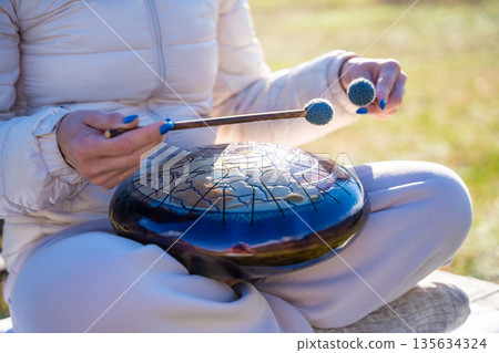 Close-up of hands playing a steel tongue drum with mallets against the backdrop of Altai mountains and river. Concept of sacred sound, touch of nature, and mindful musical ritual Close-up of hands playing a steel tongue drum with mallets against the backdrop of Altai mountains and river. Concept of sacred sound, touch of nature, and mindful musical ritual 135634324
