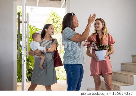 Women entering home, one holding baby, another with plant, all smiling warmly Women entering home, one holding baby, another with plant, all smiling warmly 135634383