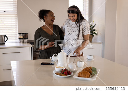 Women laughing together in kitchen, enjoying tea and pastries on counter Women laughing together in kitchen, enjoying tea and pastries on counter 135634413