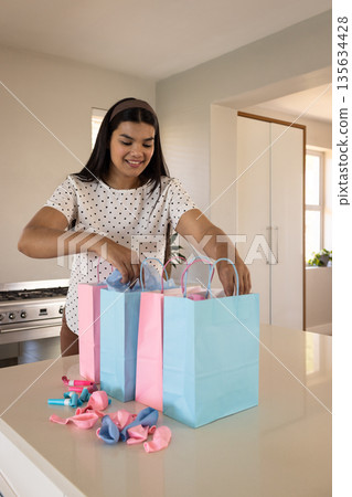 Smiling woman preparing colorful gift bags with balloons in kitchen Smiling woman preparing colorful gift bags with balloons in kitchen 135634428