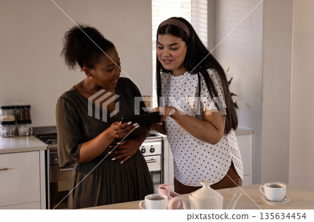 Two women in kitchen looking at tablet, discussing recipe with excitement 135634454
