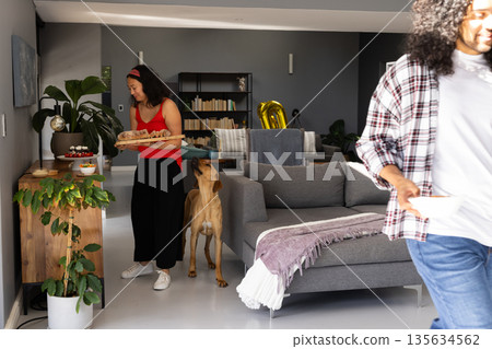 African American woman carrying tray of snacks at home, dog eagerly following 135634562