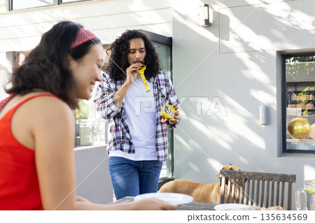 Celebrating outdoors, man blowing party horn while woman sets table with plates Celebrating outdoors, man blowing party horn while woman sets table with plates 135634569