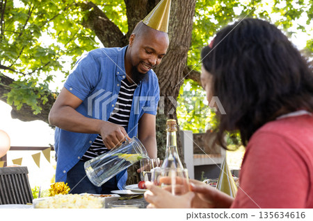 Man wearing party hat pouring lemonade at outdoor celebration with friends Man wearing party hat pouring lemonade at outdoor celebration with friends 135634616
