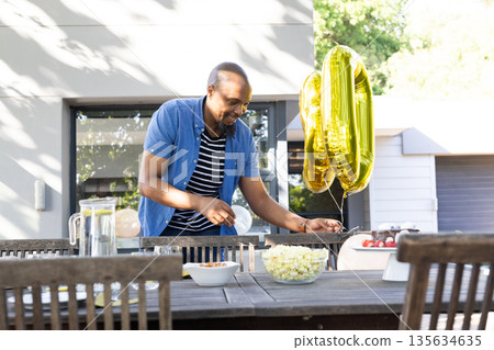 African American man arranging outdoor party table with balloons and snacks 135634635
