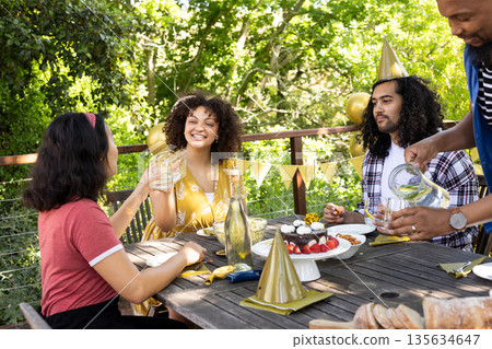 Friends celebrating birthday outdoors, enjoying food and drinks with party hats 135634647