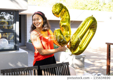 Asian woman holding gold balloons celebrating birthday outdoors, smiling joyfully 135634690