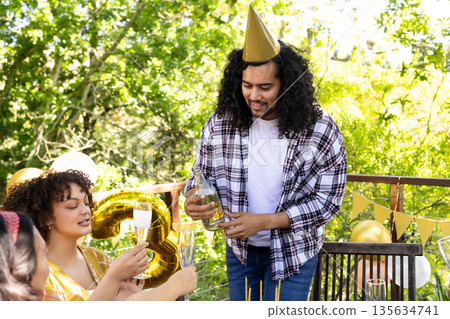 Man pouring champagne at outdoor birthday party with friends, wearing party hat Man pouring champagne at outdoor birthday party with friends, wearing party hat 135634741