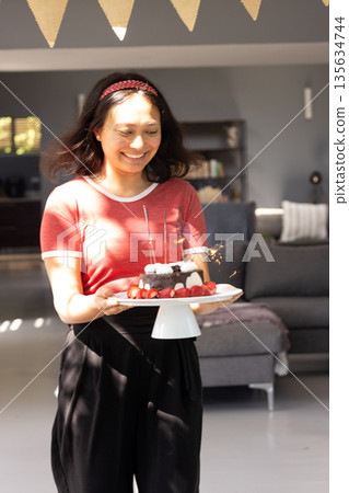 Asian woman carrying birthday cake with sparklers, smiling in living room Asian woman carrying birthday cake with sparklers, smiling in living room 135634744