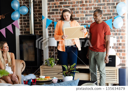Friends celebrating at home, woman holding gift while others smiling and chatting 135634879