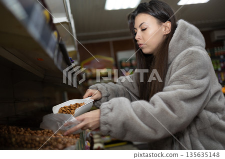 Woman in Coat Using Scoop to Put Almonds Into Plastic Bag Woman in Coat Using Scoop to Put Almonds Into Plastic Bag 135635148
