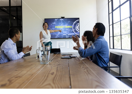 In conference room, business team applauding colleague presenting product launch on screen In conference room, business team applauding colleague presenting product launch on screen 135635347