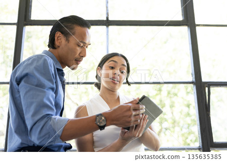 Asian man and woman discussing project using tablet in bright office setting 135635385