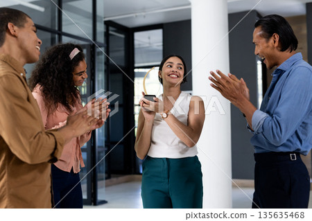 Colleagues applauding woman holding award in modern office, celebrating achievement Colleagues applauding woman holding award in modern office, celebrating achievement 135635468