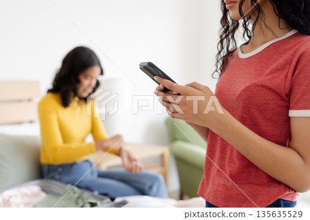 Teen using smartphone while mother sits on bed, both enjoying relaxed moment 135635529