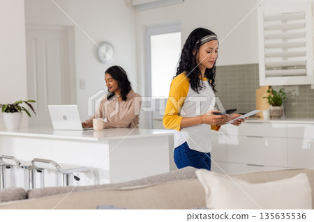 Multitasking mother using smartphone and holding paper, daughter working on laptop Multitasking mother using smartphone and holding paper, daughter working on laptop 135635536