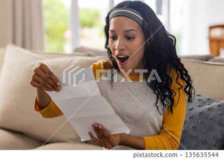 Excited woman reading letter on couch, expressing surprise and happiness at home Excited woman reading letter on couch, expressing surprise and happiness at home 135635544