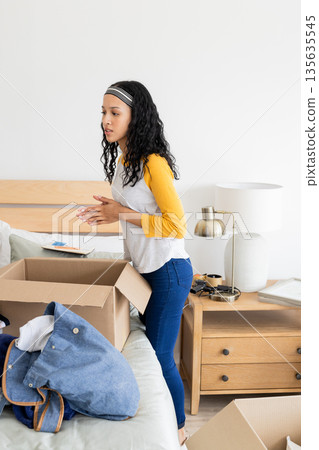 Young woman unpacking boxes in bedroom, organizing belongings with focus Young woman unpacking boxes in bedroom, organizing belongings with focus 135635545