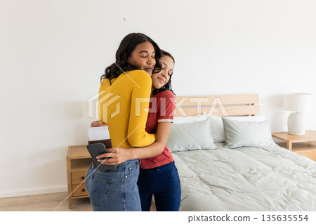 Two women hugging in bedroom, one holding passport and boarding pass, smiling Two women hugging in bedroom, one holding passport and boarding pass, smiling 135635554