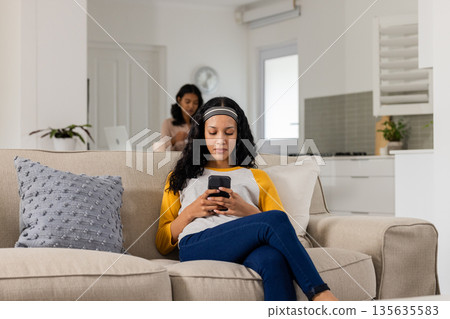 Relaxing at home, woman using smartphone while another works on laptop in background Relaxing at home, woman using smartphone while another works on laptop in background 135635583