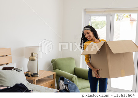 Unpacking cardboard box in bedroom, African American woman organizing belongings, smiling Unpacking cardboard box in bedroom, African American woman organizing belongings, smiling 135635604