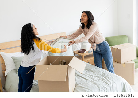 Two women unpacking boxes on bed, smiling and organizing new home together 135635613