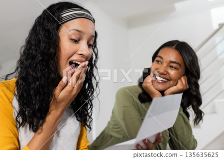 Excited African American woman reading letter while friend smiles at home Excited African American woman reading letter while friend smiles at home 135635625