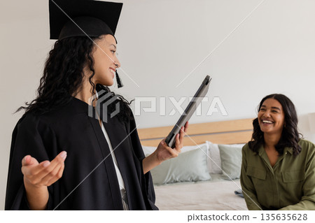 Graduation celebration, woman in cap and gown holding tablet, smiling with friend 135635628