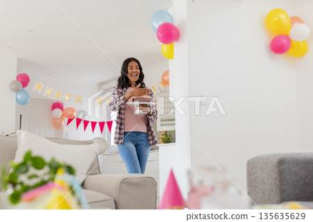 Smiling woman carrying birthday cake at home with festive decorations 135635629