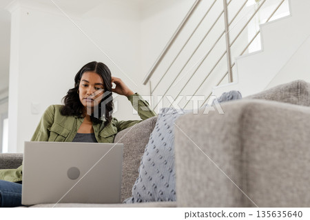 African American woman using laptop on sofa at home, looking focused and thoughtful 135635640