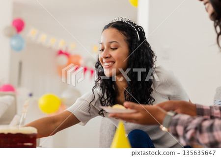 Smiling woman celebrating birthday at home, reaching for cake with friends 135635647