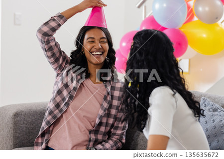Celebrating at home, joyful woman wearing party hat with friend on couch Celebrating at home, joyful woman wearing party hat with friend on couch 135635672