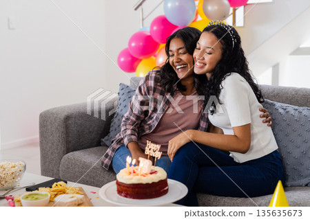 Celebrating birthday, two women hugging on couch with cake and balloons 135635673