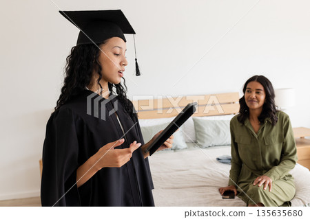 Asian graduate in cap and gown reading speech at home with proud mother watching Asian graduate in cap and gown reading speech at home with proud mother watching 135635680