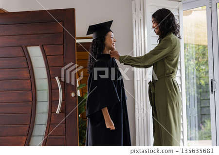 Mother adjusting daughter's graduation gown at home, both smiling proudly 135635681
