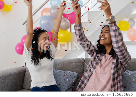 Celebrating at home, two women joyfully throwing confetti with colorful balloons 135635690