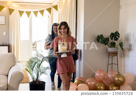 Smiling woman carrying birthday cake with candles, preparing surprise party at home Smiling woman carrying birthday cake with candles, preparing surprise party at home 135635728