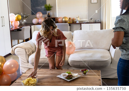 Woman arranging snacks on table in living room, preparing for celebration Woman arranging snacks on table in living room, preparing for celebration 135635730