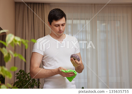 Man cleaning house, carefully wiping a decorative object in his living room 135635755