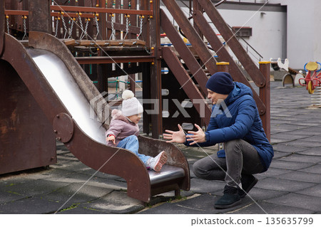 Father catching his toddler daughter sliding down a playground slide 135635799
